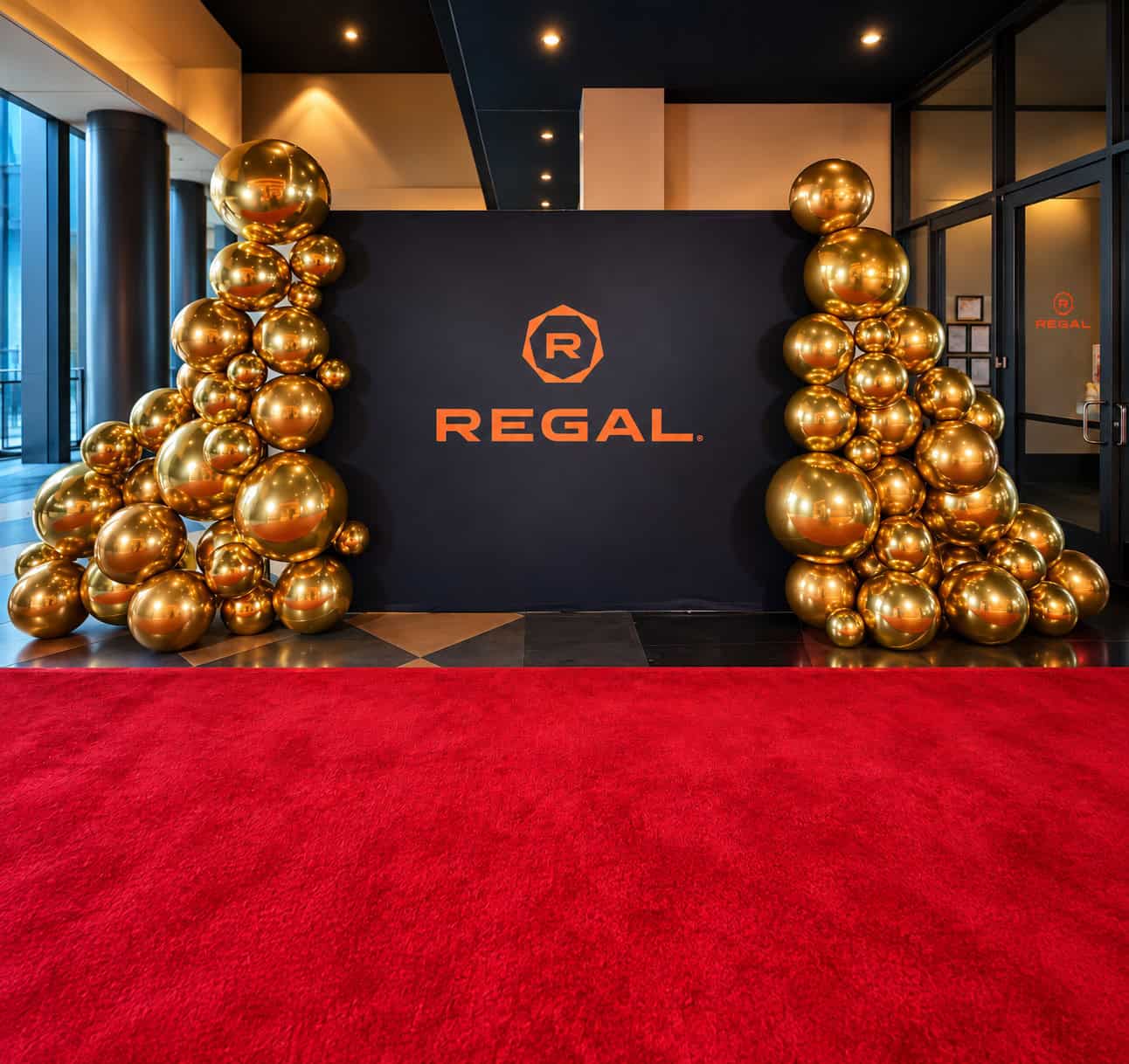 A Regal-branded photo backdrop flanked by custom balloon garlands in shimmering gold stands behind a red carpet in a modern, well-lit lobby.