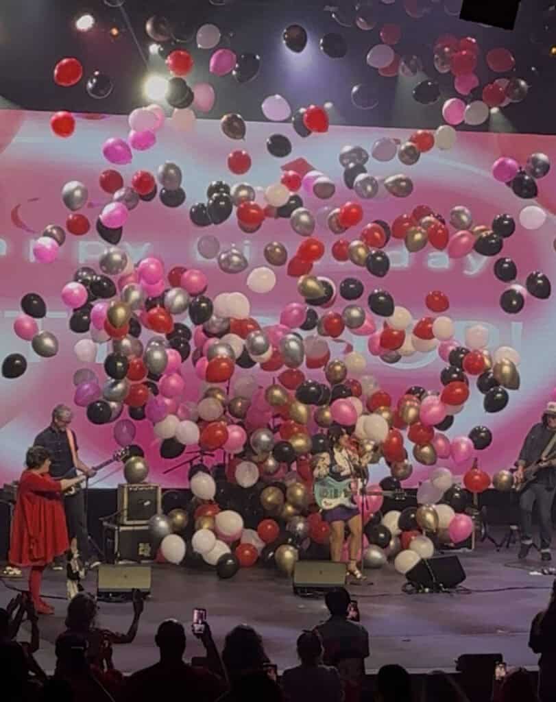 A band performs on stage while red, white, pink, and black balloon drops fill the air from above, with audience members taking photos and a large illuminated screen in the background.