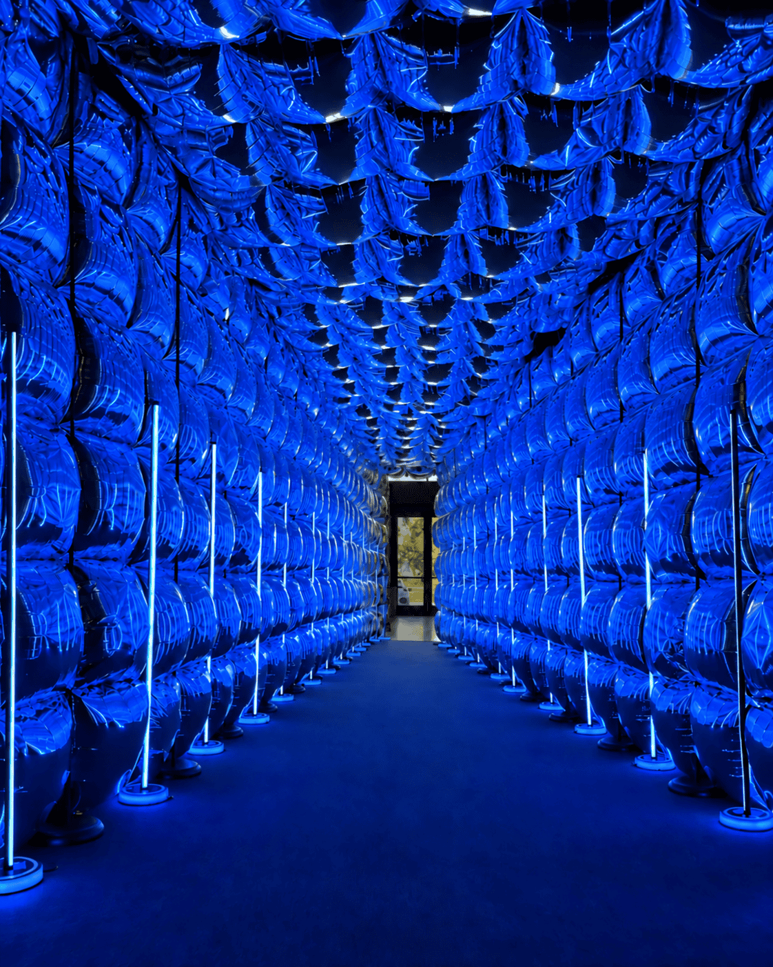 A hallway lined with rows of reflective, blue-lit inflated panels, creating a tunnel-like effect leading to an open doorway.