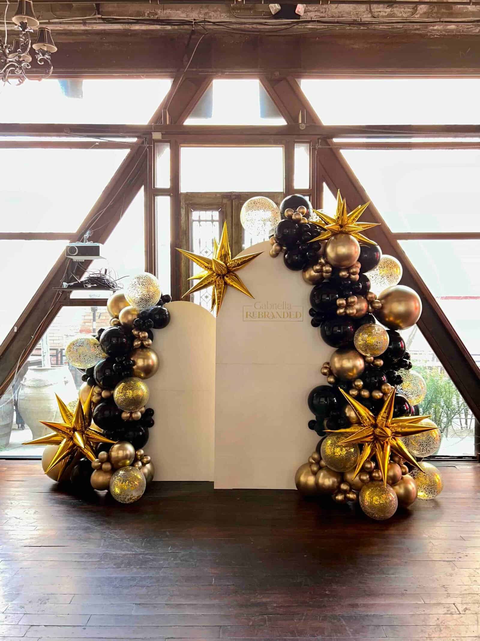 Black, gold, and white balloon arch frames a white backdrop. A starburst decorates each side.