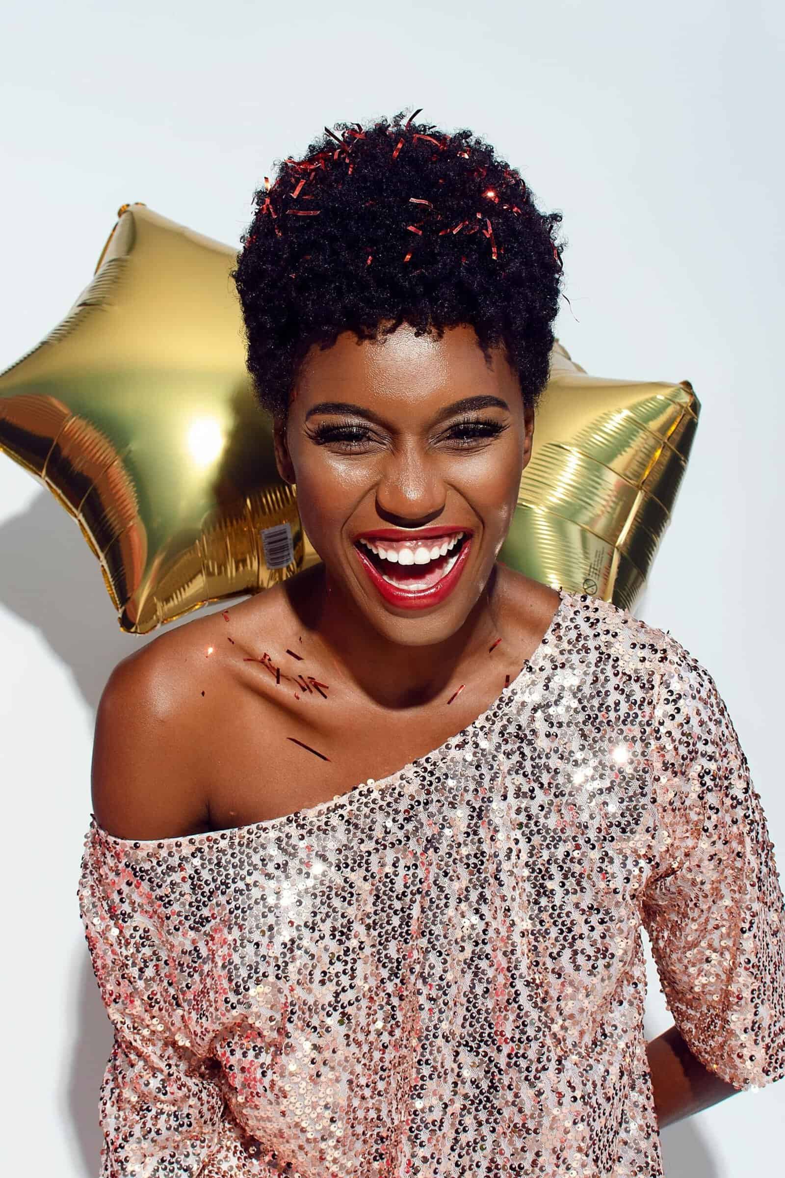A woman in a sequined top is smiling while holding two gold balloons.
